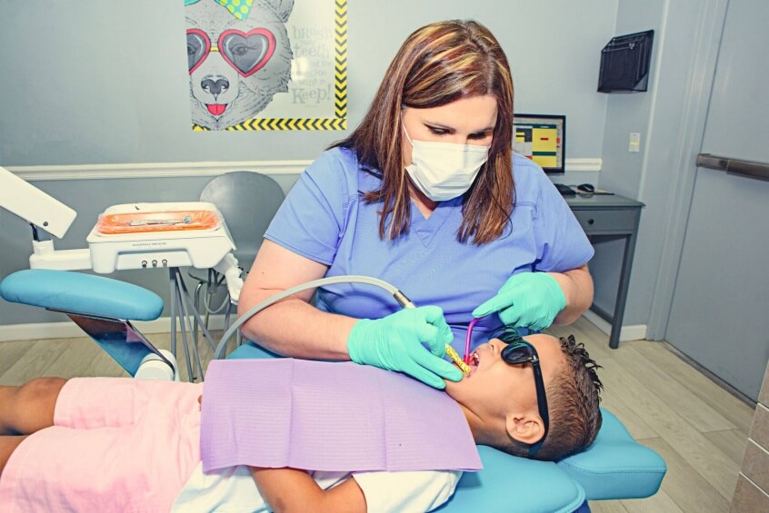 Route 32 Dental team member wearing a mouth and nose mask is working with suction tools cleaning inside the mouth of a young boy patient lying on the dentist treatment chair wearing protective dark glasses