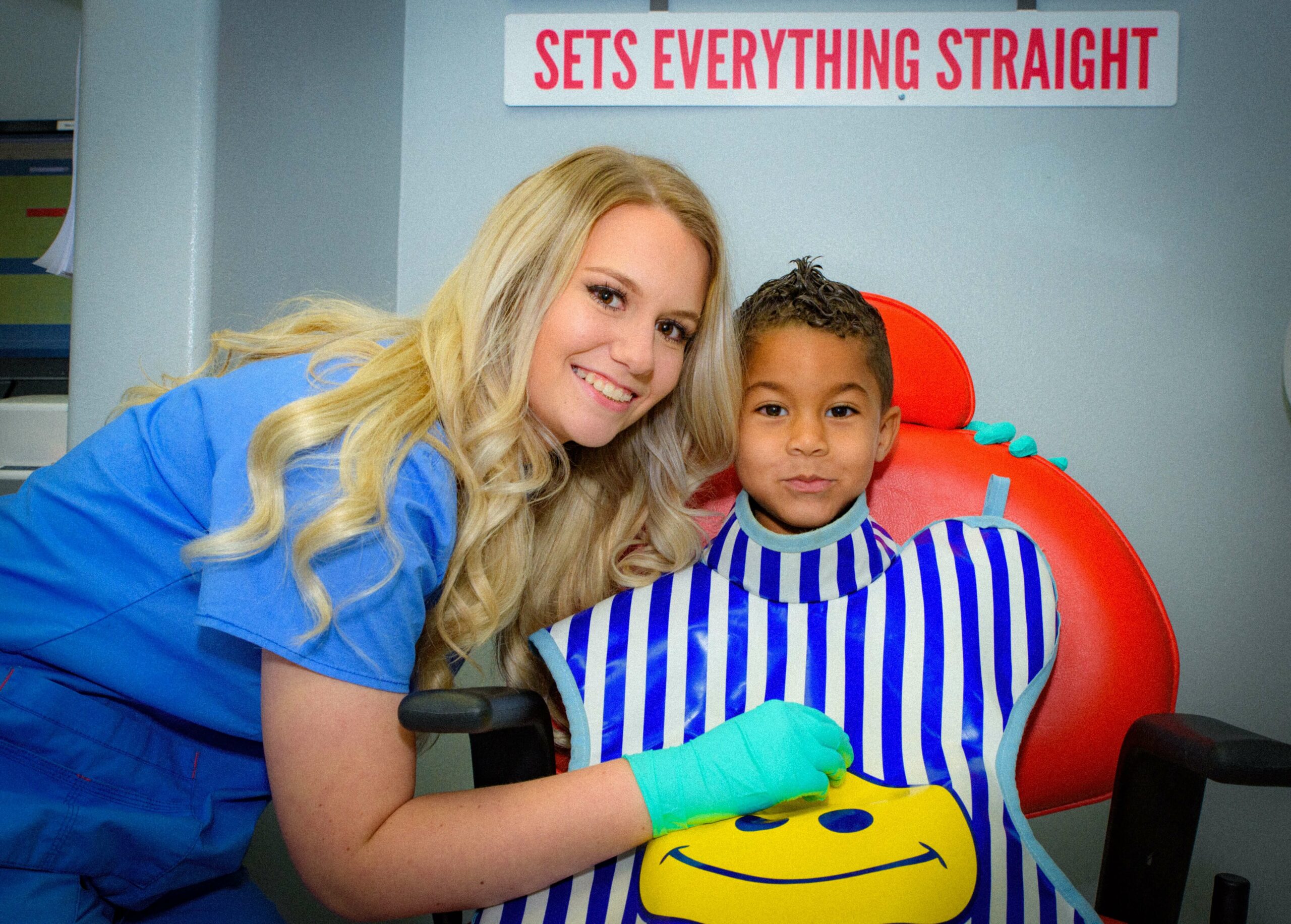 A smiling Route 32 Dental team member wearing office uniform and a young patient wearing the protective X-Ray apron with blue stripes and a smiley face print, sitting on a red dentist chair