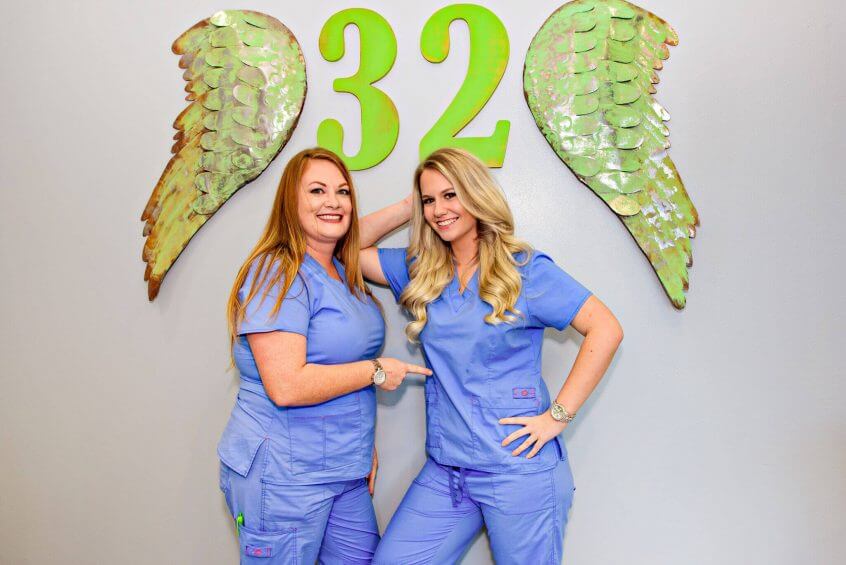Two Route 32 Dental team members in office uniforms posing under the angel wings 32 sign at the office
